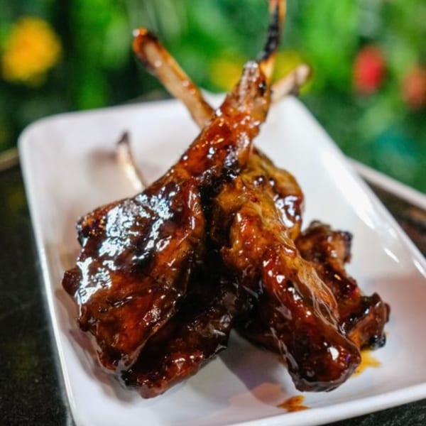 Glazed lamb lollipops on a white rectangular plate, with a green and yellow blurred background.