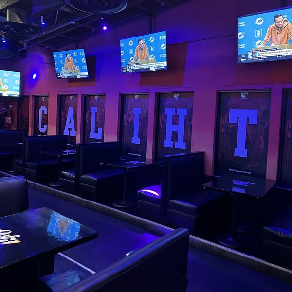 Dark Cali Kitchen and Bar interior with booths, tables, and TVs displaying sports, illuminated by purple lighting.