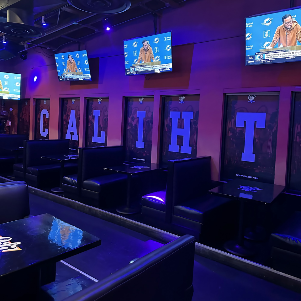 Dark Cali Kitchen and Bar interior with booths, tables, and TVs displaying sports, illuminated by purple lighting.