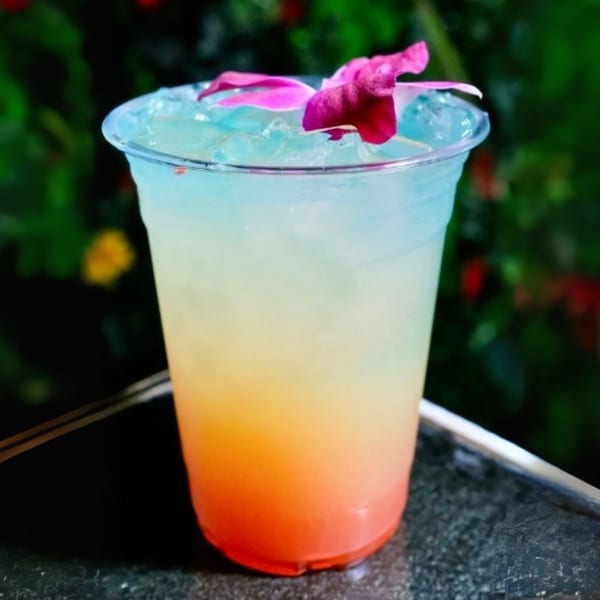 A vibrant, layered cocktail with blue, yellow, and red hues, garnished with a pink orchid.